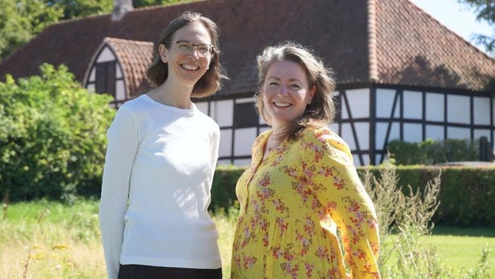 To glade kvinder poser uden for et charmerende bindingsværkshus i en frodig grøn omgivelse.