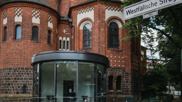 Das Bild zeigt ein modernes Glasgebäude vor einem historischen Backsteingebäude mit einem Straßenschild 