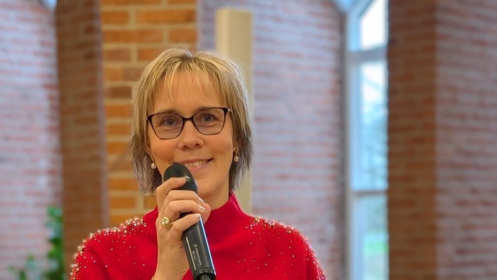 Kvinde i rød trøje og sort skørt står ved pult og holder mikrofon.