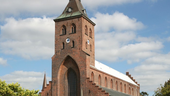 Historisk murstenskirke med højt tårn og buede vinduer under en delvist overskyet himmel.