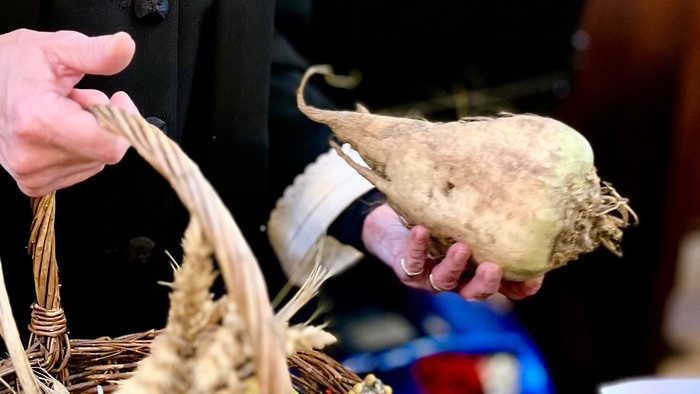 En person i formelt tøj holder en vævet kurv med en udstoppet fugl og blomster i et indendørs miljø.