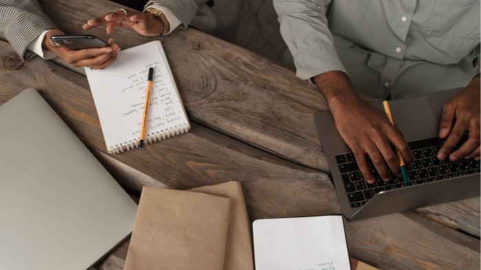 Træbord det ovenfra med mapper, papir, noteblok og blyant.  2 personer sidder ved siden af hinanden deres hænder kan ses. En taster på en bærbar med pen i hånden den anden holder en telefon 