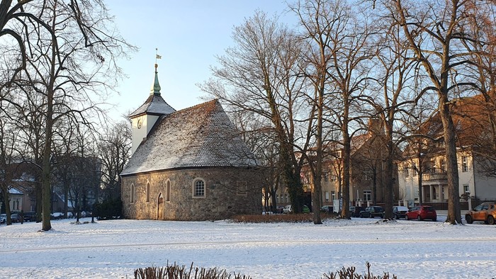 Schneelandschaft mit alter Steinkirche, kahlen Bäumen und umliegenden Gebäuden.