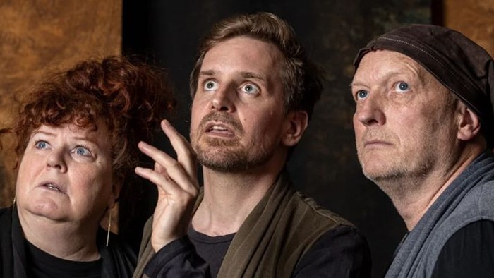 Tre skuespillere med intense udtryk poser tæt sammen i en dramatisk, tekstureret kulisse.