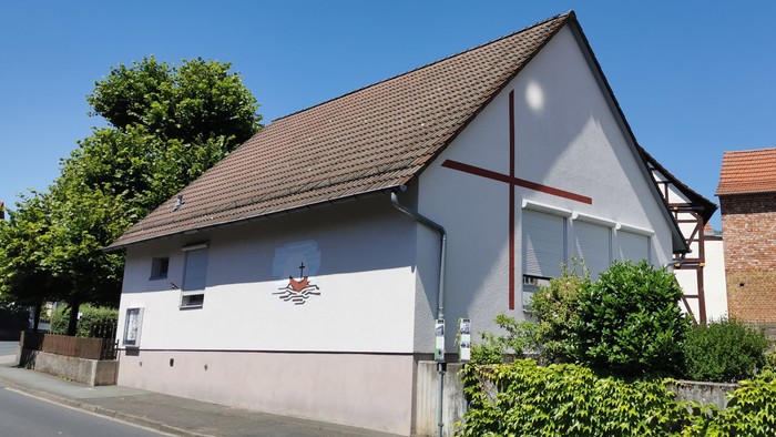 Kleine Kirche mit Kreuz an der Seite, umgeben von Büschen und Bäumen, neben einem Ziegelgebäude.