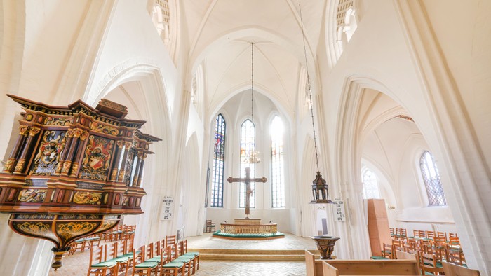Elegant kirkeinteriør med udsmykket prædikestol, træbænke og buede farvede glasvinduer under blødt lys.