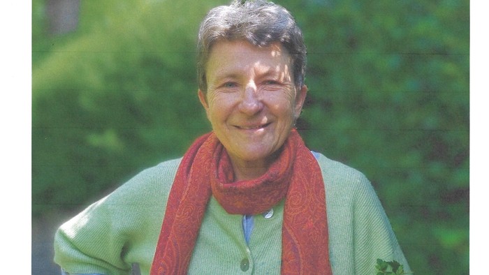 En glad person iført en grøn jakke og et rødt tørklæde udendørs med grønne omgivelser i baggrunden.