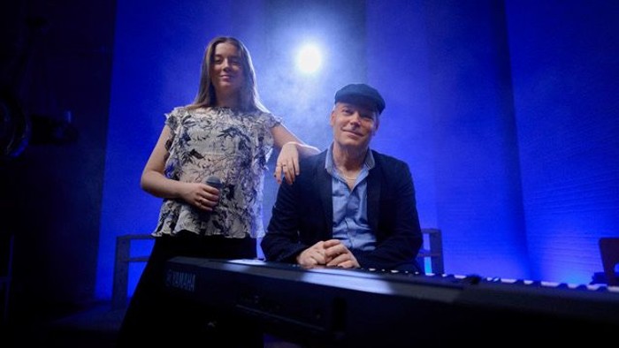 Foto af Timeless duo: Monica Rosenberg stående og Ken Thinggaard siddende vad klaver, i modlys med blå baggrund