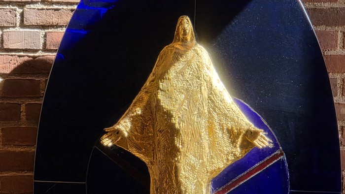 Forgyldt statue af en klædt figur oplyst mod en mørk, reflekterende cirkulær baggrund.