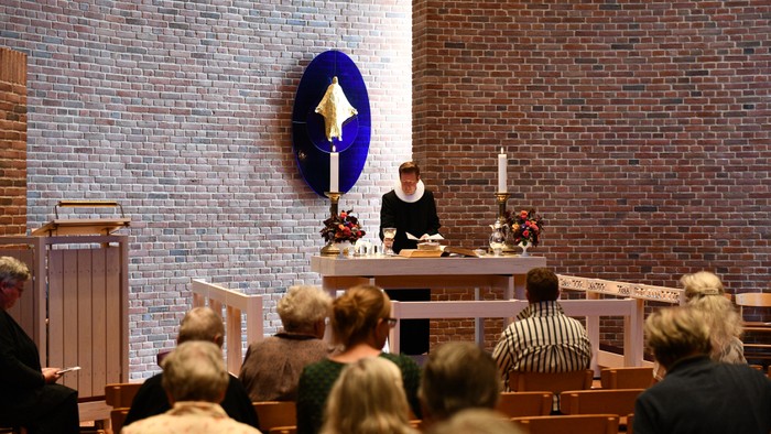 En højtidelig gudstjeneste med en præst, der leder de deltagende, som sidder i kirkebænke, og lys tændt på alteret.