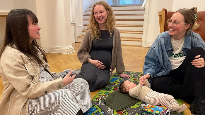 Tre kvinder sidder på gulvmatte, hvor en af dem holder et barn. De ser lykkelige ud og er optaget af hinanden.