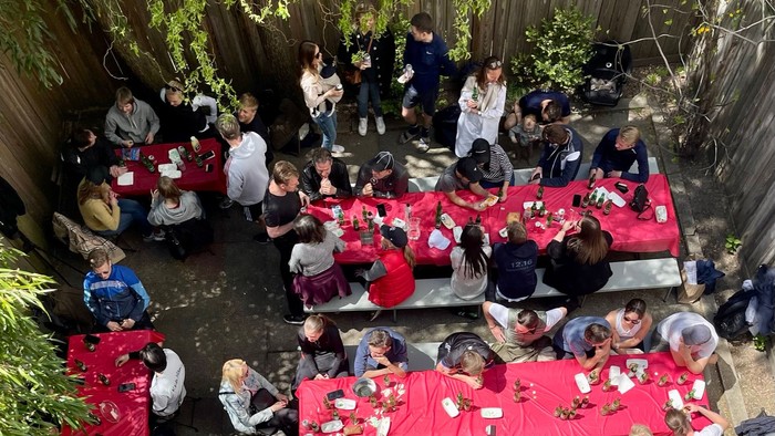 En stor gruppe af mennesker sidder ved lange bord dækket med røde dækkinger i en have.