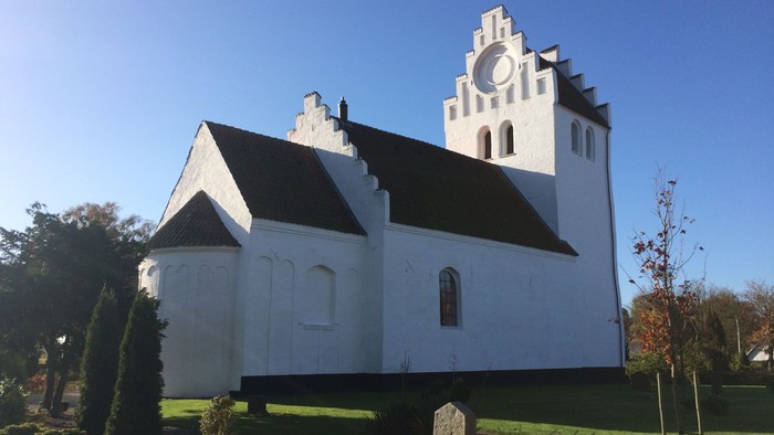 En hvid kirke med sort tag og tårn, omgivet af græs og træer