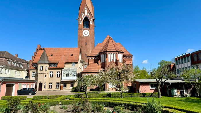 Bild zeigt eine Kirche mit hohem Turm und Uhr, umgeben von einem gepflegten Garten.