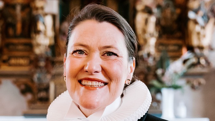 En kvinde i sort jakke med hvid krage står foran en prægtig altertavle.