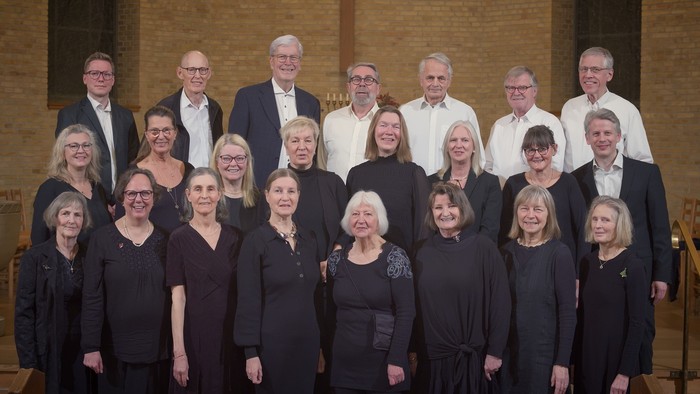 Et formelt gruppefoto af et kor med mænd i jakkesæt og kvinder i sort påklædning, der poser sammen.