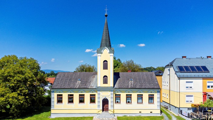 Kirche mit hohem Turm und gelbem Gebäude, umgeben von grünem Gras und blauem Himmel.