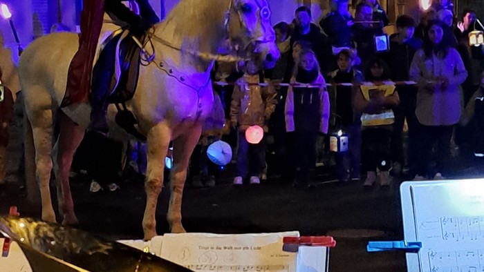 Eine nächtliche Parade-Szene mit einem Reiter zu Pferd, Musikern, die Blechblasinstrumente spielen, und einer Menschenmenge im Hintergrund.