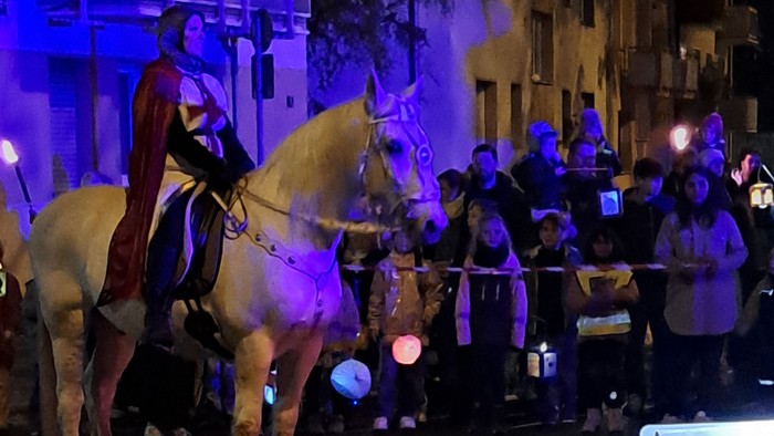 Eine nächtliche Parade-Szene mit einem Reiter zu Pferd, Musikern, die Blechblasinstrumente spielen, und einer Menschenmenge im Hintergrund.