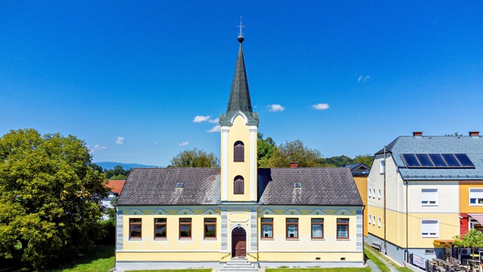 Kirche mit hohem Turm und gelbem Gebäude, umgeben von grünem Gras und blauem Himmel.