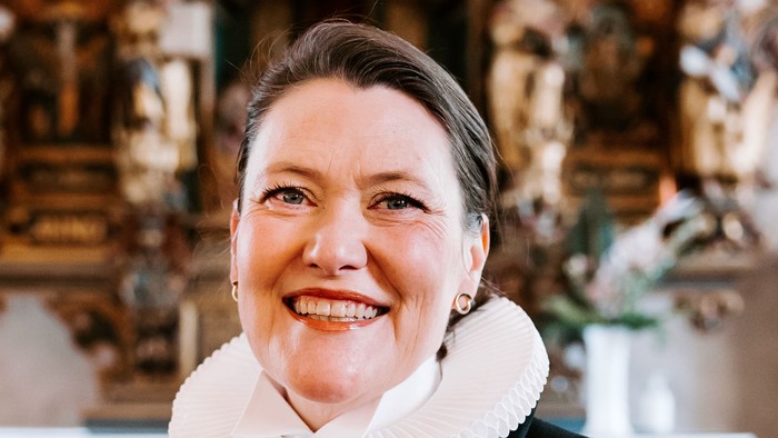 En kvinde i sort jakke med hvid krage står foran en prægtig altertavle.