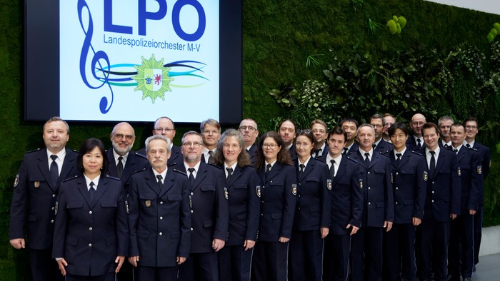 Ein großer Chor posiert vor einem grünen Hintergrund mit einem LPO-Logo.