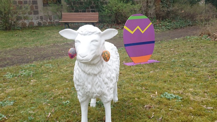 Weiße Schafskulptur vor buntem Ei und Steinmauer