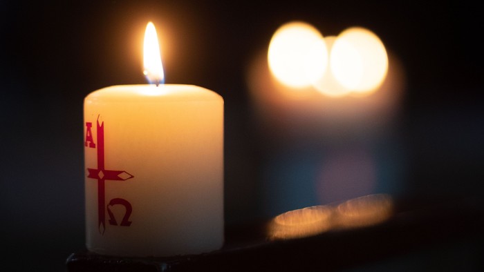 Eine brennende Kerze mit Kreuzsymbol im Vordergrund, zwei weitere Kerzen im Hintergrund