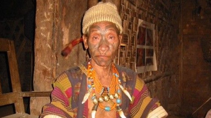 En person iført farverigt traditionelt tøj sidder indendørs med en pibe og bærer dekorative benbånd.