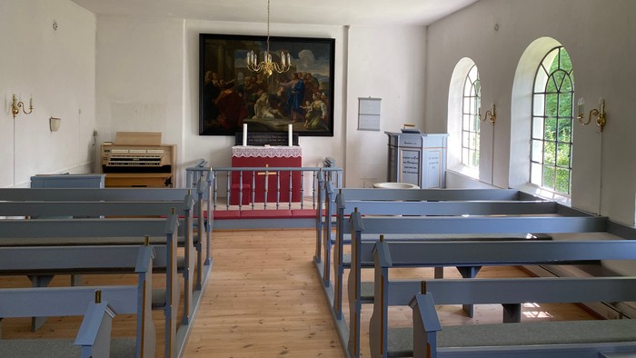 Hyggelig kirkeindretning med træbænke, alter og et stort religiøst maleri over prædikestolen.