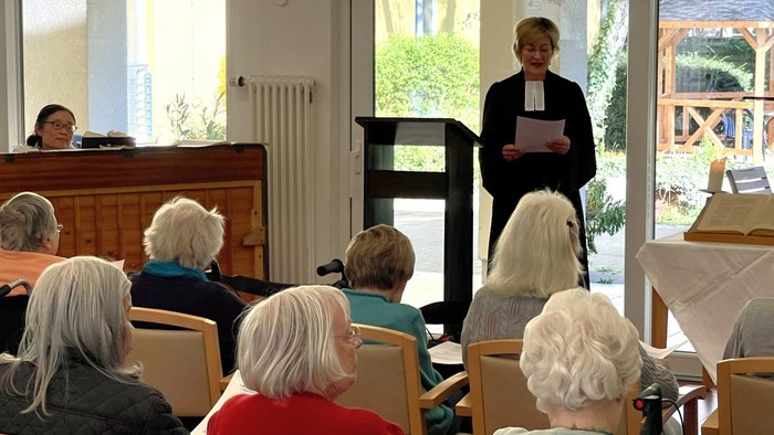 Eine Frau hält eine Rede vor einer Gruppe älterer Menschen in einem Raum mit großen Fenstern.