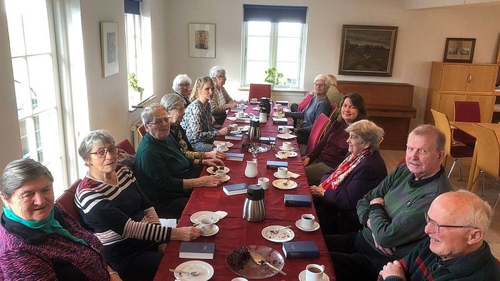 En gruppe nyder te og samtale ved et langt fællesbord i et hyggeligt lokale.