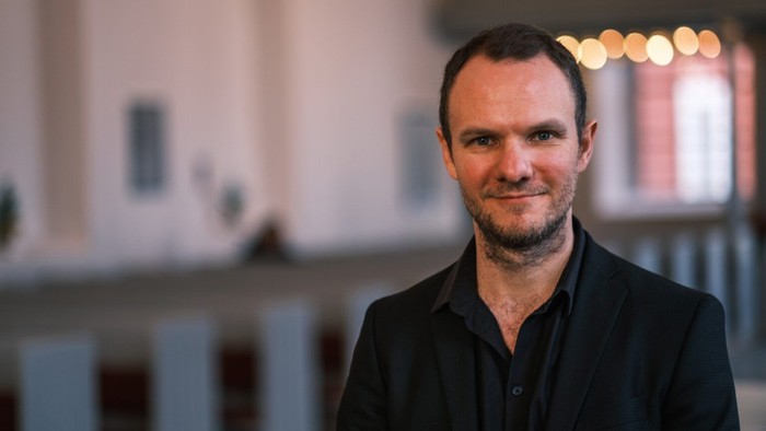 En mand i sort jakkesæt smiler indendørs nær en stilfuld, svagt oplyst arkitektonisk baggrund.