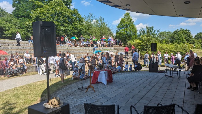 Open-Air-Veranstaltung mit sitzenden Gästen, die unter einem Pavillon in einer Parkanlage Musik und Essen genießen.