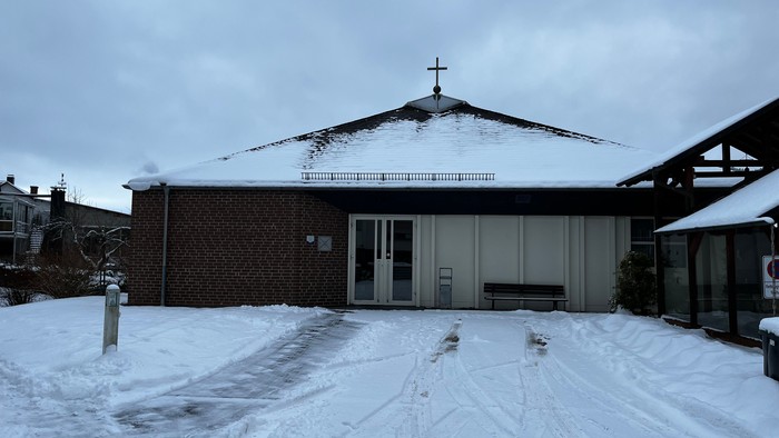Schneebedecktes Gebäude mit einem Kreuz auf der Spitze, umgeben von einem leicht eingeschneiten Parkplatz.