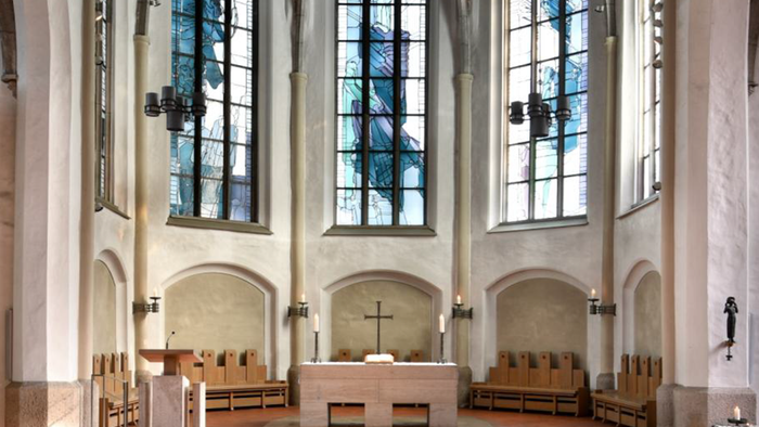 Moderne Kircheninnenraum mit hohen Buntglasfenstern, hölzernen Kirchenbänken und einem schlichten Altar mit einem Kreuz.