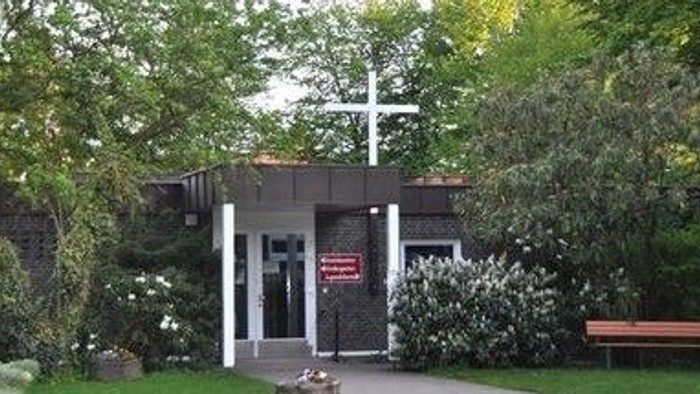 Eine Kirche mit einem Kreuz und einem Schild, umgeben von Bäumen und Sträuchern, mit einer Bank in der Nähe.