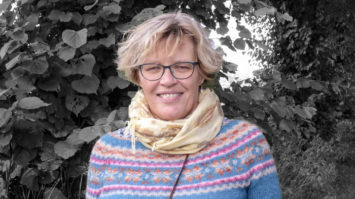 En person med briller, en farverig sweater og et tørklæde står på en løvfyldt, snoet landevej.