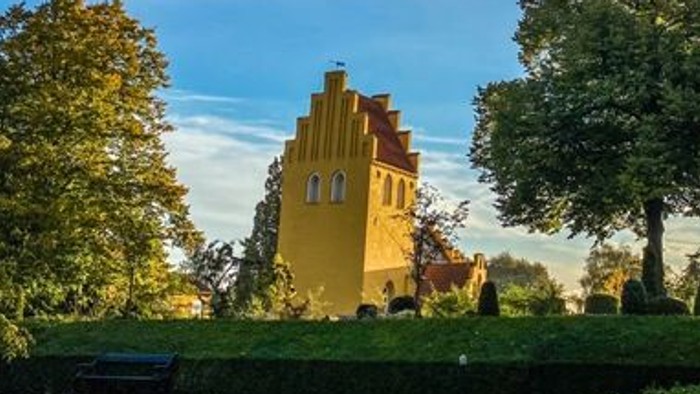 En malerisk gul kirke står omgivet af frodige grønne træer under en klar, levende himmel.