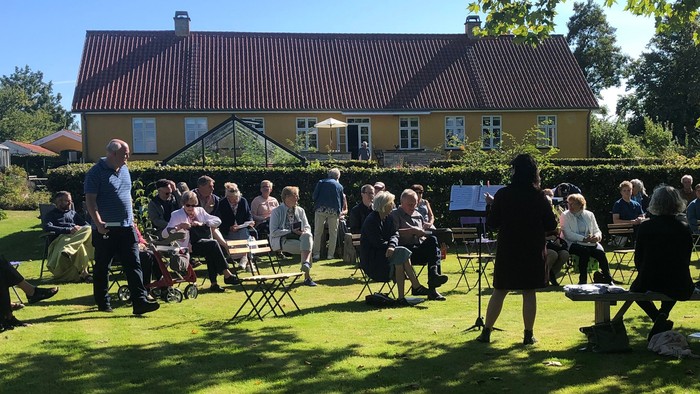 En gruppe af mennesker sidder ude på græs og holder fest ved et hus.