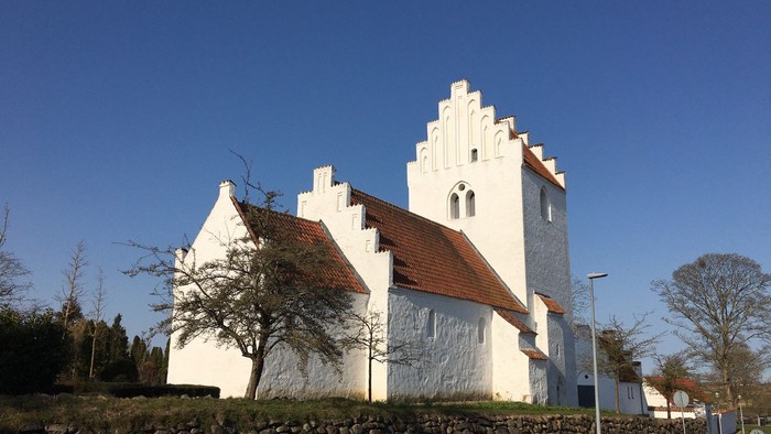 Et hvidt kirkebygning med spistegnet tak og tårn, omgivet af en mur og træer