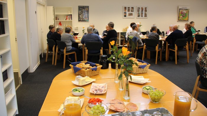 I forgrunden et ta´-selv-bord dækket op med juice, frugt, pålæg, ost og boller. I baggrunden ældre mennesker omkring nogle borde.