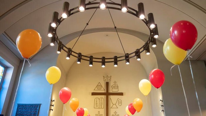 Bunte Luftballons schmücken das Innere einer Kirche unter einer beleuchteten, runden Lichtinstallation und einem Kruzifix.