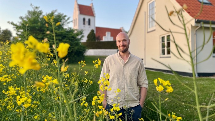 En mand står midt i levende gule blomster i en frodig have med et idyllisk hus i baggrunden.