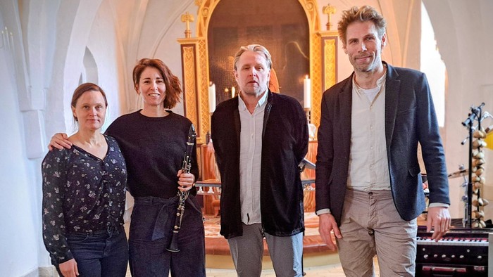 En gruppe på fire musikere, der poser sammen inde i en historisk kirke med musikudstyr i nærheden.