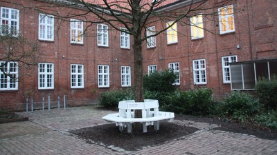 Im Innenhof eines Ziegelgebäudes steht eine weiße Bank unter einem Baum.