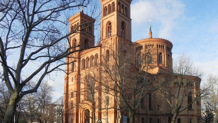 Großes, altes Backsteingebäude mit zwei Türmen und Rundbau, umgeben von kahlen Bäumen und einem Auto