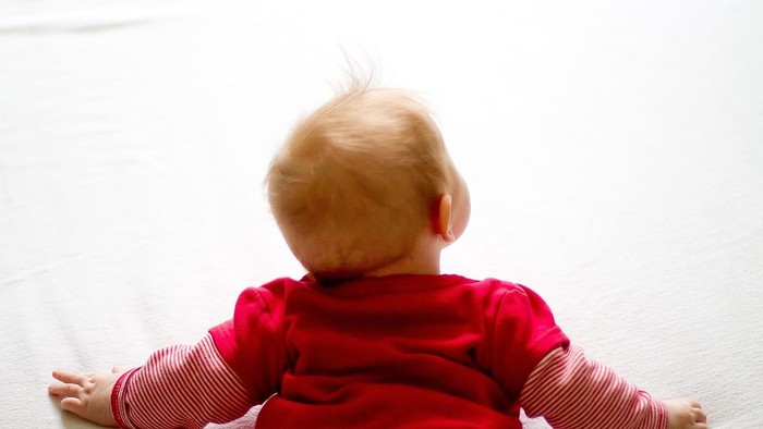 Baby liegt auf dem Rücken in einem rot-rosa gestreiften Strampler auf einer weichen Unterlage.