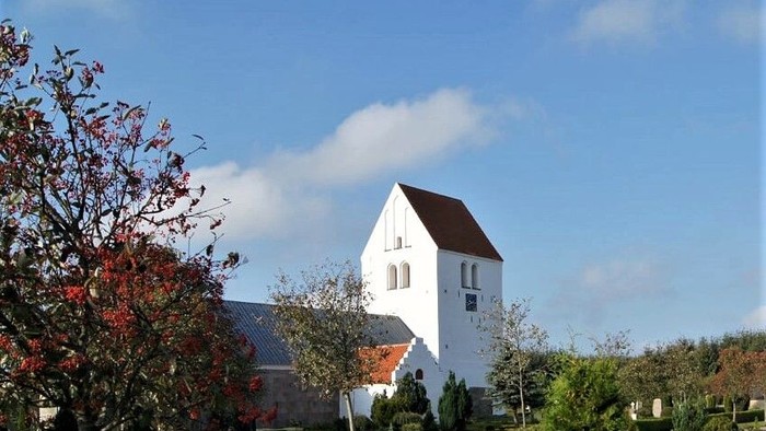 En hyggelig hvid kirke med et højt spir mod en klar blå himmel og efterårsløv.