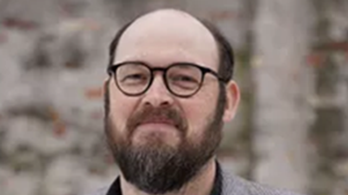 En skægget mand med briller iført en grå jakke på en struktureret baggrund.